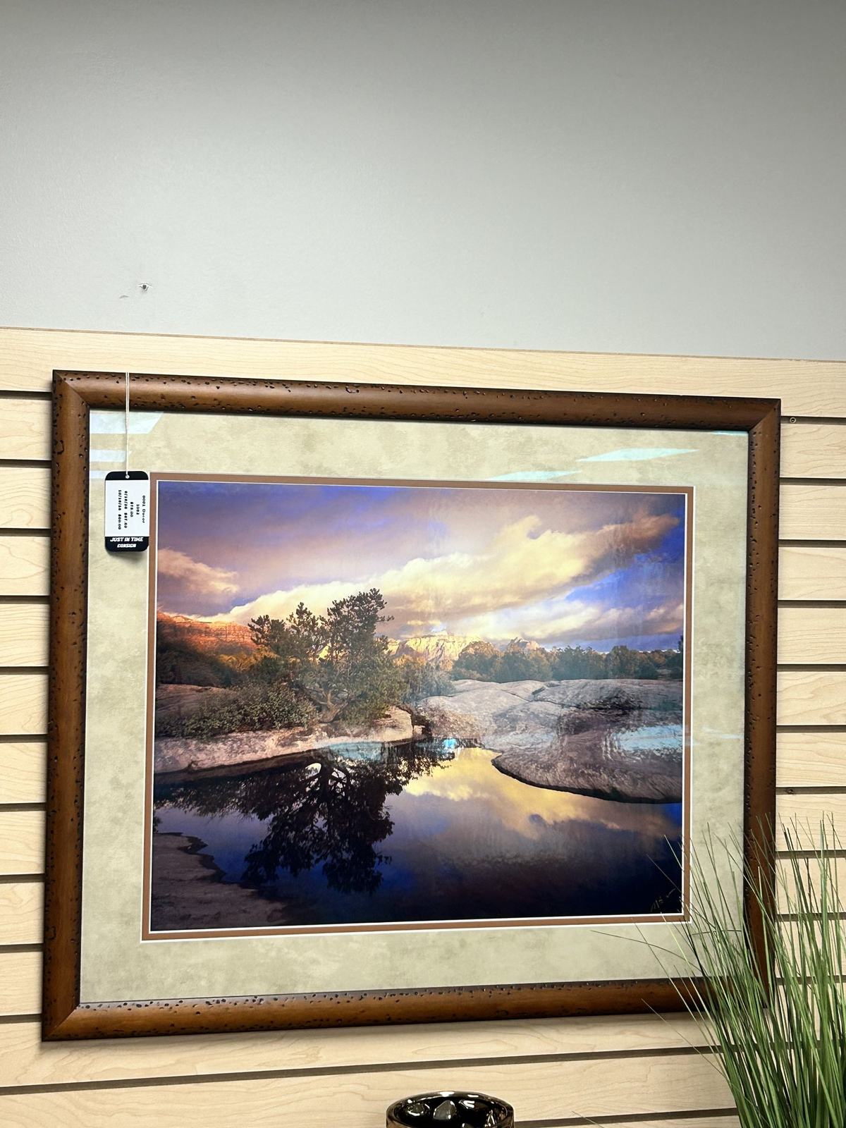 A framed landscape painting of a river canyon at sunset, displayed on slatwall