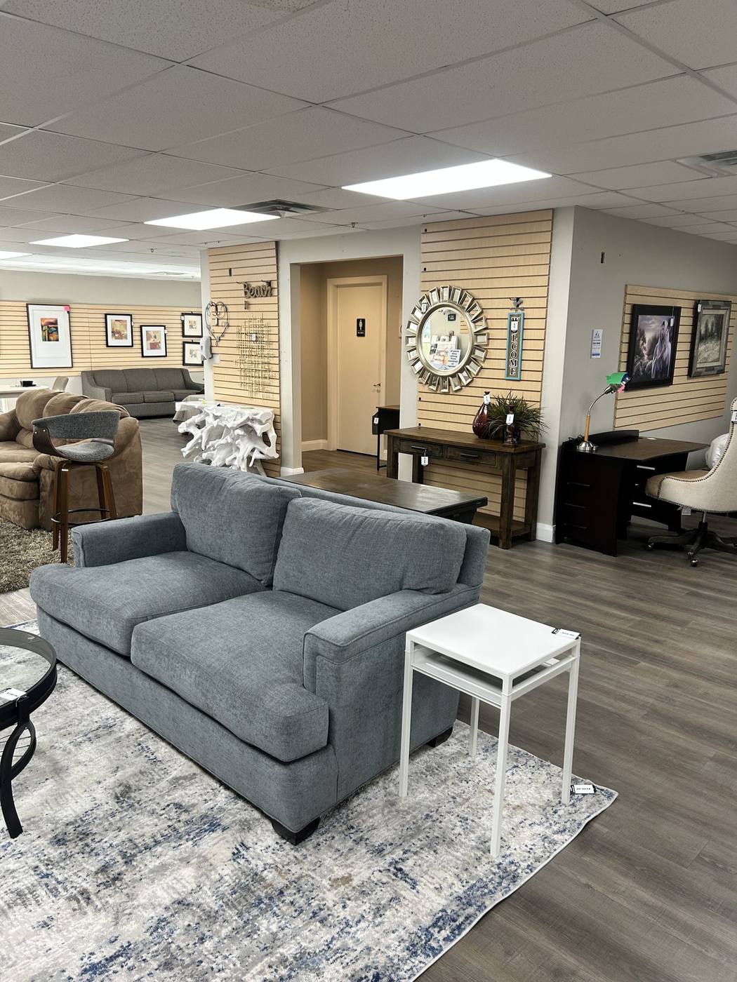 A grey two-cushion sofa styled with a patterned rug and white side table on the consignment showroom floor