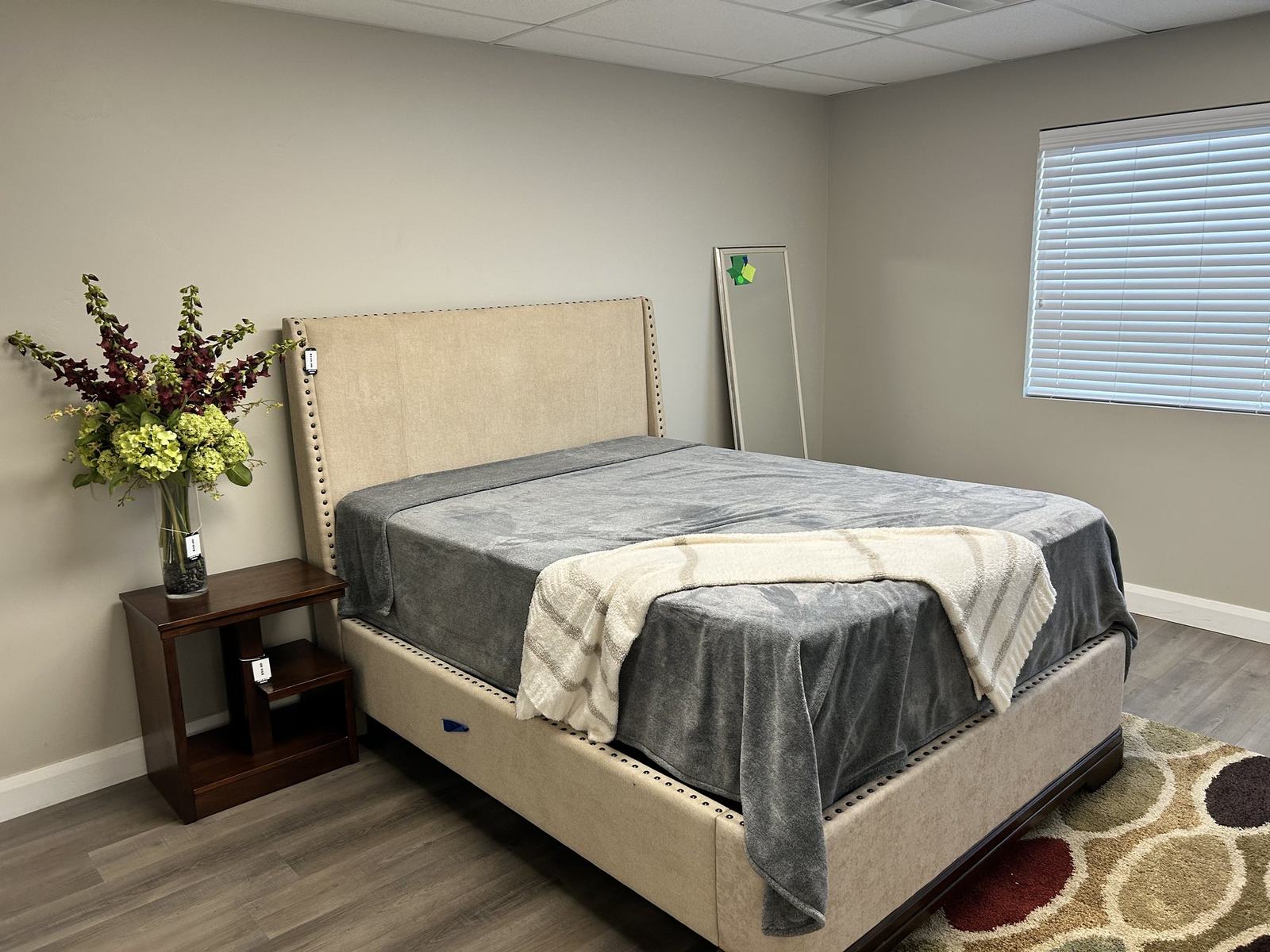 An upholstered bed with a grey throw, wooden nightstand and fresh flowers in the consignment showroom